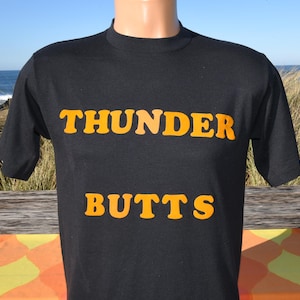 May include: Black t-shirt with the words "THUNDER BUTTS" in orange block letters. The shirt is short-sleeved and has a crew neck. The background shows a beach scene with the ocean and some grass.