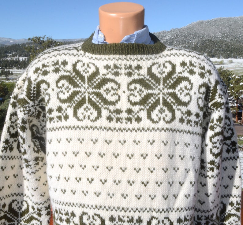 May include: A white and green knitted jumper with a traditional Scandinavian pattern. The jumper has a crew neck and long sleeves.