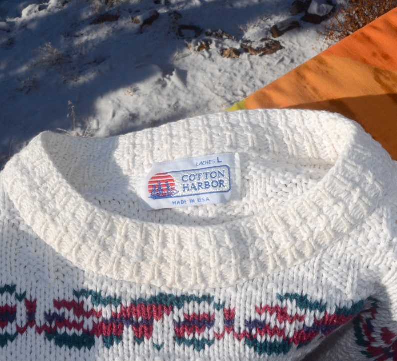 May include: White cotton sweater with a red, green, and blue geometric pattern. The sweater has a crew neck and a label that reads "Ladies L Cotton Harbor Made in USA."
