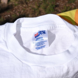 以下が含まれることがあります： 「Pre-shrunk Pre-retreci Pre-encogido」と「100% Cotton Coton Algodon Fabrique aux E.U. Hecho en los E.U.A. Made in U.S.A.」と「Grand/Grande Large (14-16)」のテキストが書かれた、白い Hanes Beefy-T シャツのタグ