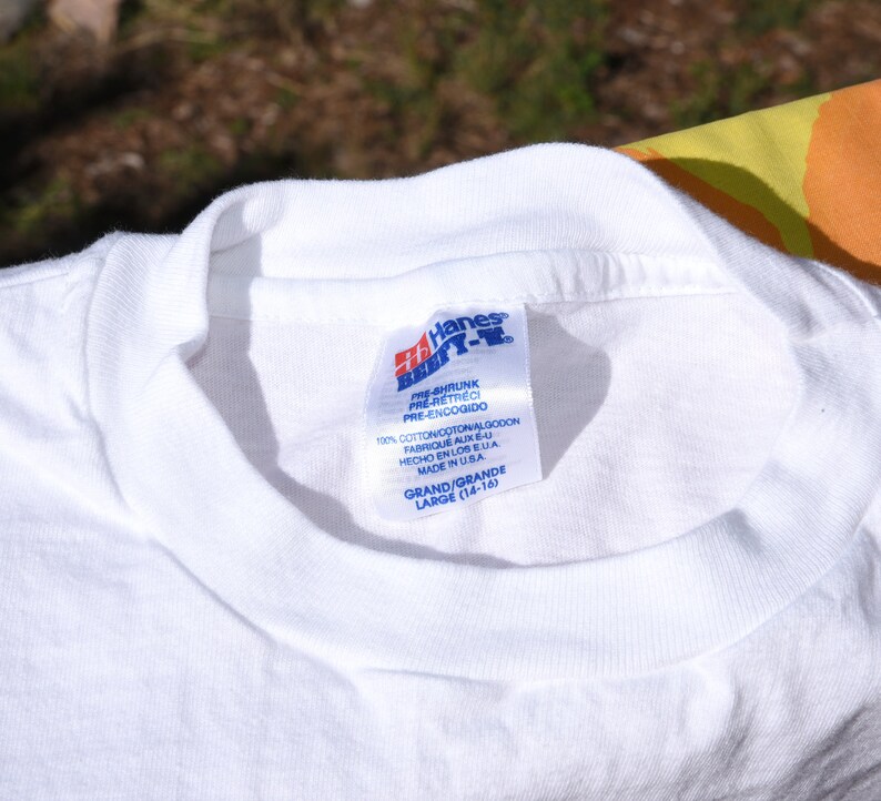 以下が含まれることがあります： 「Pre-shrunk Pre-retreci Pre-encogido」と「100% Cotton Coton Algodon Fabrique aux E.U. Hecho en los E.U.A. Made in U.S.A.」と「Grand/Grande Large (14-16)」のテキストが書かれた、白い Hanes Beefy-T シャツのタグ