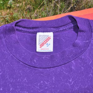 Puede incluir: Primer plano de una camiseta morada con cuello redondo y un patr&oacute;n texturizado. La etiqueta dice "JERZEES" e indica que la camiseta es talla M, fabricada en EE. UU. y es 100% algod&oacute;n.