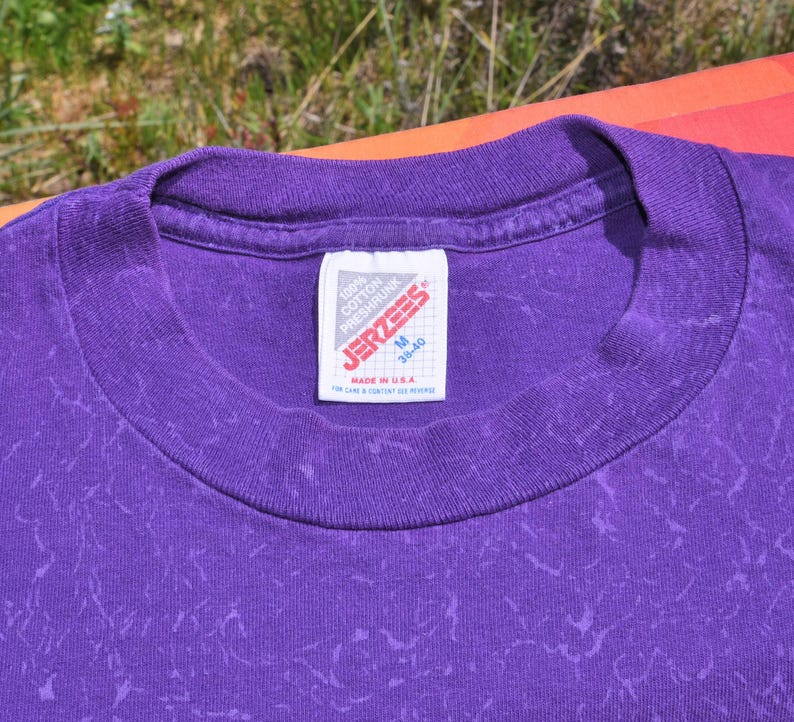 Puede incluir: Primer plano de una camiseta morada con cuello redondo y un patr&oacute;n texturizado. La etiqueta dice "JERZEES" e indica que la camiseta es talla M, fabricada en EE. UU. y es 100% algod&oacute;n.