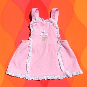 Puede incluir: Un vestido de beb&eacute; rosa con ribete de encaje blanco y flores bordadas. El vestido tiene tirantes y un cierre de bot&oacute;n en la espalda.