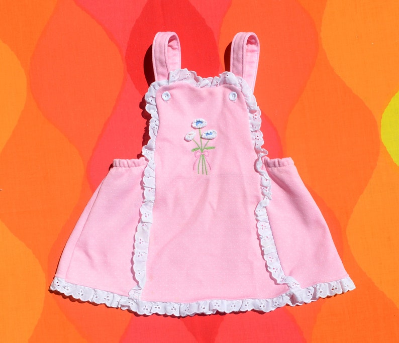 Puede incluir: Un vestido de beb&eacute; rosa con ribete de encaje blanco y flores bordadas. El vestido tiene tirantes y un cierre de bot&oacute;n en la espalda.