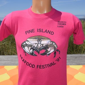 May include: A bright pink short-sleeve t-shirt featuring a graphic of a crab in the center. The text reads PINE ISLAND SEAFOOD FESTIVAL '91 in black lettering, with ORGANIZED FISHERMEN OF FLORIDA printed in smaller black text near the shoulder.