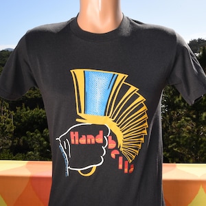 May include: A white ceramic mug with a black handle and the text "But First, Coffee" in a black, handwritten-style font. The mug is on a white surface.