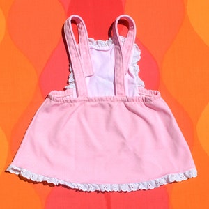 Puede incluir: Un vestido rosa sin mangas con ribete de encaje blanco alrededor del escote y el dobladillo. El vestido tiene dos tirantes que van sobre los hombros.