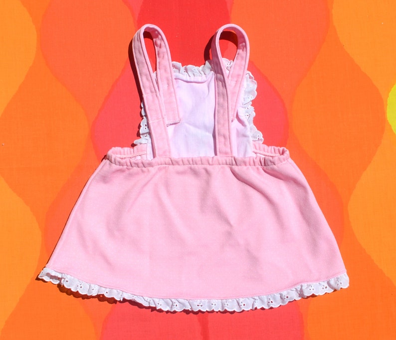 Puede incluir: Un vestido rosa sin mangas con ribete de encaje blanco alrededor del escote y el dobladillo. El vestido tiene dos tirantes que van sobre los hombros.