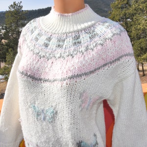 May include: White knitted sweater with a pink, gray, and blue geometric pattern around the neck and butterfly designs throughout.