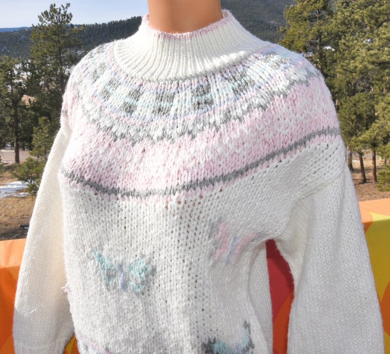 May include: White knitted sweater with a pink, gray, and blue geometric pattern around the neck and butterfly designs throughout.