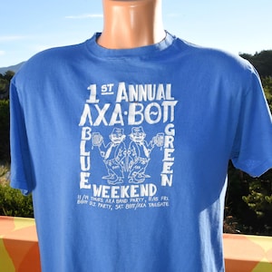 May include: A blue t-shirt with white text and graphics. The text reads "1st Annual AXA BOTT Weekend" with additional text below. The graphic features two cartoon figures holding mugs. The shirt is displayed on a mannequin.