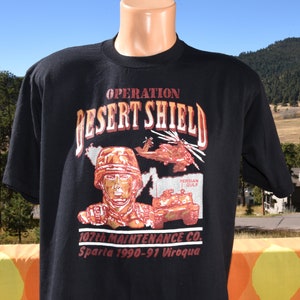 May include: Black t-shirt with a graphic print of a soldier, a helicopter, and a tank. The text "Operation Desert Shield" is printed in red and orange. The text "107th Maintenance Co. Sparta 1990-91 Viroqua" is printed below the graphic.