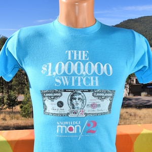 May include: A light blue t-shirt with the text "THE $1,000,000 SWITCH" in white. A mock-up of a $1,000,000 bill is printed on the shirt with the text "KNOWLEDGE man/2" below it.