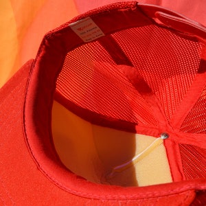 Op de afbeelding: Rode mesh trucker cap met een wit label met de tekst "100% Polyester, Maat One Size Fits All, Gemaakt in Korea."