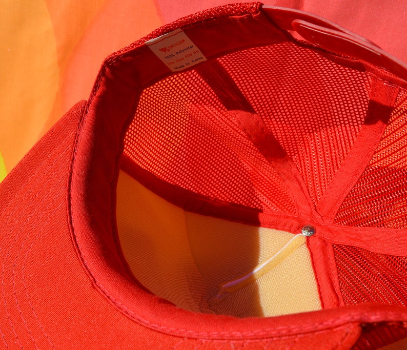 Op de afbeelding: Rode mesh trucker cap met een wit label met de tekst "100% Polyester, Maat One Size Fits All, Gemaakt in Korea."