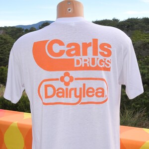 May include: White t-shirt with orange text. The shirt features the words "Carls DRUGS" in a bold, stylized font above the word "DairyLea" in a rounded rectangle with a flower graphic. The shirt is displayed on a mannequin.