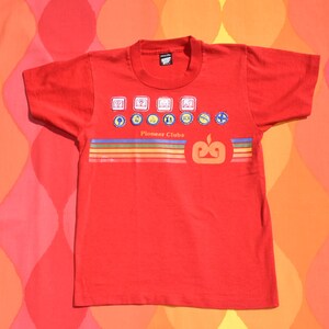 May include: A red t-shirt featuring the text "Pioneer Clubs" with various colorful graphic icons and a stylized apple design. The shirt has a crew neck and short sleeves, perfect for casual wear.