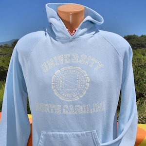 以下が含まれることがあります： 白文字で「UNIVERSITY OF NORTH CAROLINA CHAPEL HILL」と書かれた水色のパーカー。パーカーには、ドローストリング付きのフード、フロントポケット、長袖が付いています。大学の紋章も表示されています。