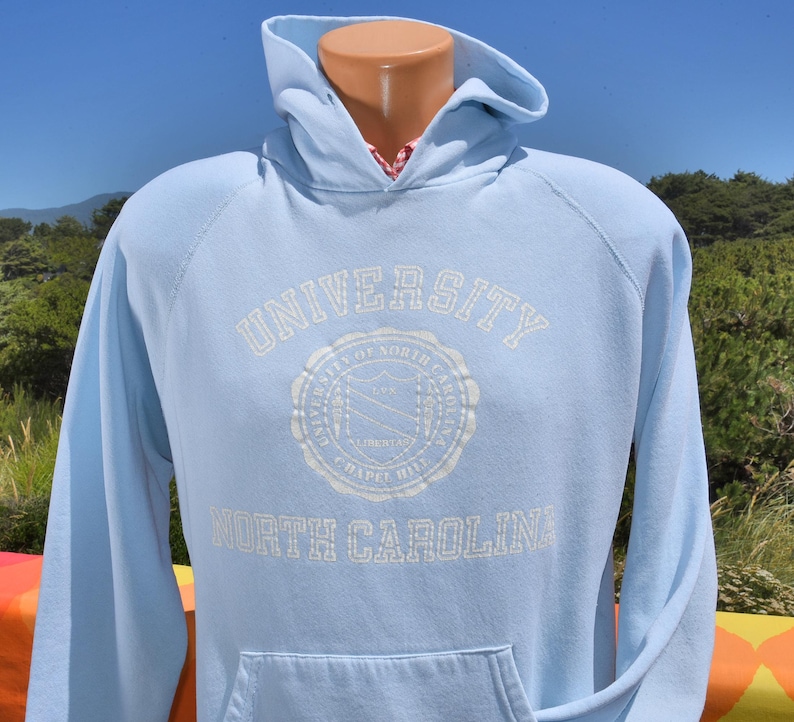 以下が含まれることがあります： 白文字で「UNIVERSITY OF NORTH CAROLINA CHAPEL HILL」と書かれた水色のパーカー。パーカーには、ドローストリング付きのフード、フロントポケット、長袖が付いています。大学の紋章も表示されています。