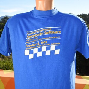 May include: A royal blue t-shirt with white text that reads "Creative Think Presents Success In Software" and additional text below. The shirt also features a checkered design. The text includes the date October 4, 1983, and the location Hyatt Rickeys Hotel, Palo Alto, California.