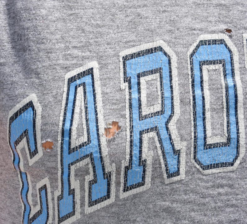 May include: Close-up of a gray sweatshirt with the word "CAROLINA" in large, distressed, light blue letters outlined in white. The fabric shows signs of wear and tear, with small holes visible.
