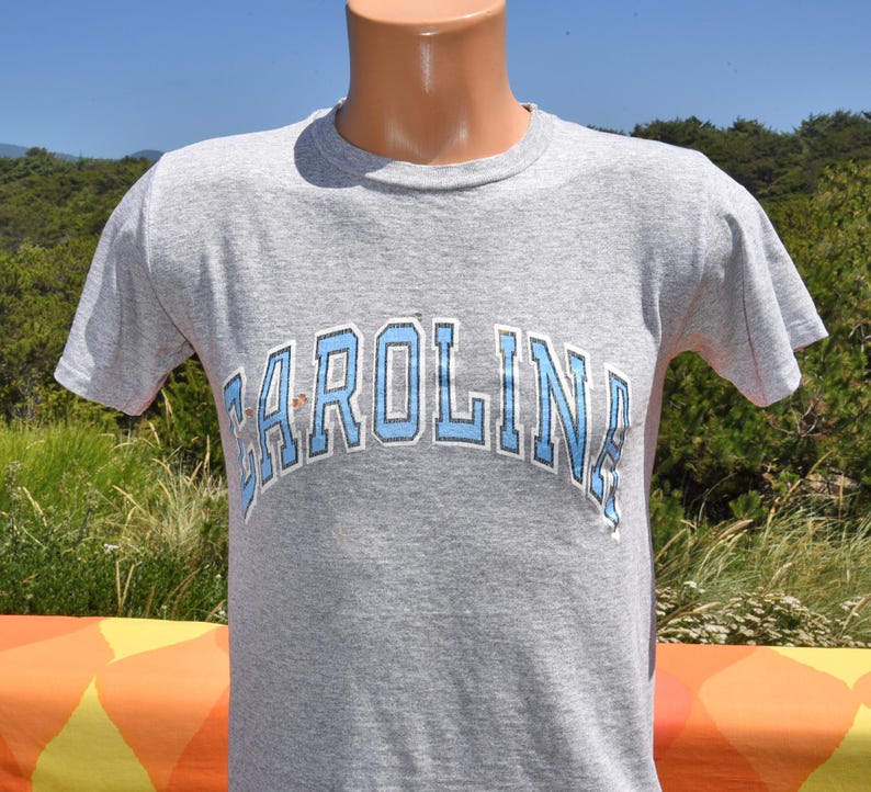 May include: A gray t-shirt with the word "CAROLINA" arched across the chest in light blue and white lettering. The shirt has short sleeves and a crew neck. The shirt is displayed on a mannequin.