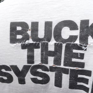 Op de afbeelding: Wit T-shirt met de tekst "BUCK THE SYSTEM" gedrukt in zwarte letters. De letters zijn licht beschadigd en hebben zichtbare naden.