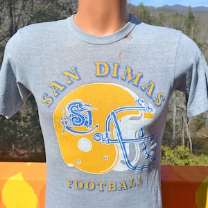 May include: A gray t-shirt featuring a yellow and blue football helmet graphic with the words "SAN DIMAS FOOTBALL". The shirt has short sleeves and a crew neck.