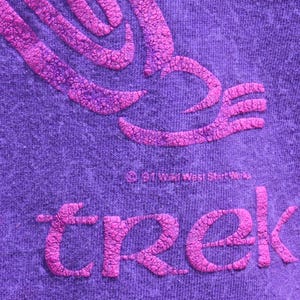 Puede incluir: Primer plano de una tela morada con un dise&ntilde;o rosa en relieve y texturizado. La palabra "trek" se muestra en rosa, junto con un gr&aacute;fico estilizado. La tela tiene un aspecto jaspeado.