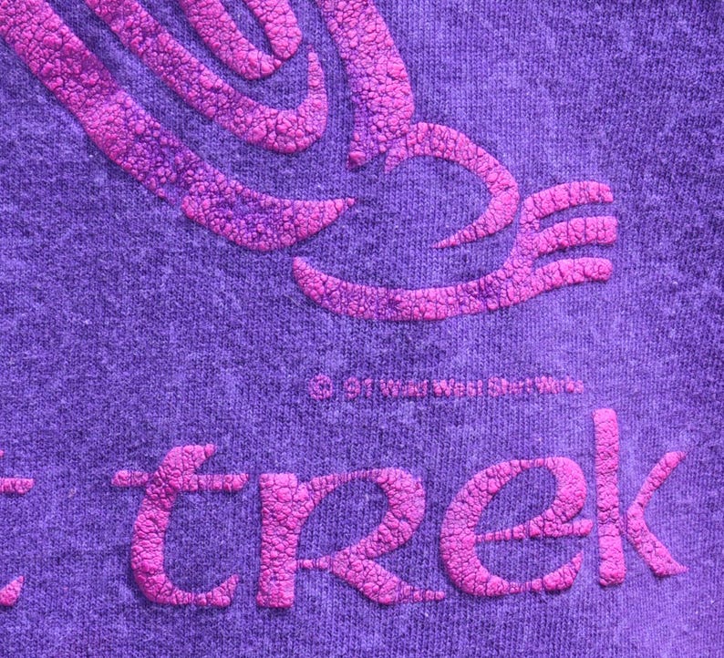 Puede incluir: Primer plano de una tela morada con un dise&ntilde;o rosa en relieve y texturizado. La palabra "trek" se muestra en rosa, junto con un gr&aacute;fico estilizado. La tela tiene un aspecto jaspeado.