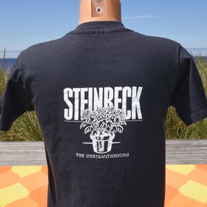 May include: A black short-sleeved t-shirt featuring a white graphic print on the back. The design displays the word STEINBECK in large capital letters above a line drawing of a potted plant, with the text THE CHRYSANTHEMUMS printed below it.