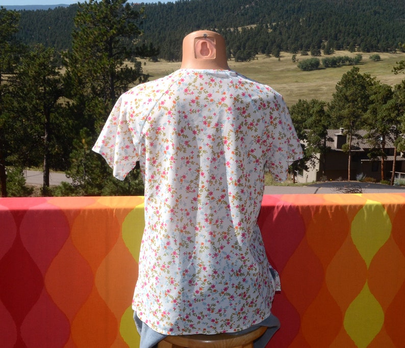 May include: White short-sleeved shirt with a floral print in shades of pink, blue, and orange. The shirt has a round neckline and a relaxed fit.