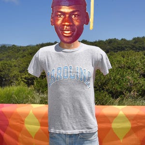 May include: A gray t-shirt with the word "Carolina" in light blue arched across the chest. The shirt is worn by a figure with a cutout of a person's face and a graduation cap. The background features a colorful table and green foliage.
