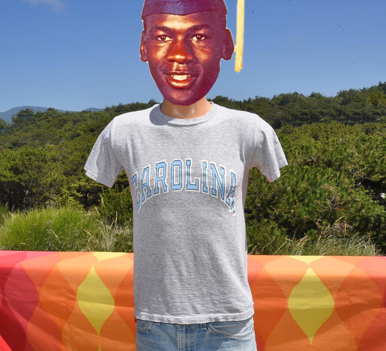 May include: A gray t-shirt with the word "Carolina" in light blue arched across the chest. The shirt is worn by a figure with a cutout of a person's face and a graduation cap. The background features a colorful table and green foliage.