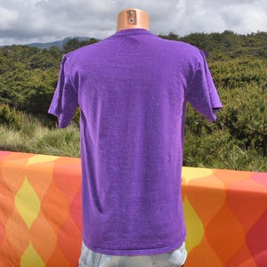 Puede incluir: Una camiseta morada con apariencia texturizada se exhibe en un maniqu&iacute;. La camiseta de manga corta est&aacute; hecha de un material suave. El fondo presenta una superficie estampada colorida y un entorno natural al aire libre.