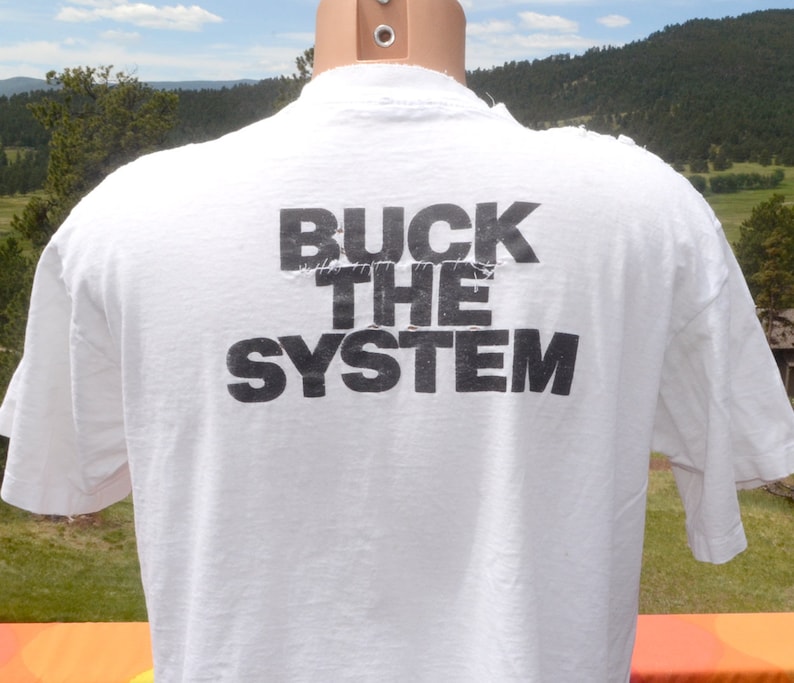 Op de afbeelding: Wit T-shirt met de tekst "BUCK THE SYSTEM" in zwart gedrukt op de achterkant.
