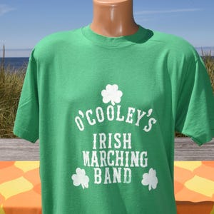 May include: A bright green short-sleeved t-shirt featuring a white graphic print. The design includes a shamrock icon above the text O'COOLEY'S IRISH MARCHING BAND, with two additional shamrocks positioned below the text in a distressed white font.