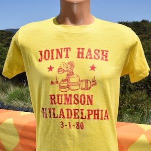 May include: Yellow t-shirt with red text and graphic. The text reads "JOINT HASH RUMSON HILADELPHIA 3-1-80". The graphic features a cartoon character and barrels. The shirt is a crew neck with short sleeves.