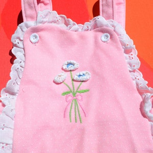 Puede incluir: Mono rosa para beb&eacute; con ribete de encaje blanco y flores bordadas. Las flores son blancas con centros azules y tallos verdes. Hay un lazo rosa en los tallos.