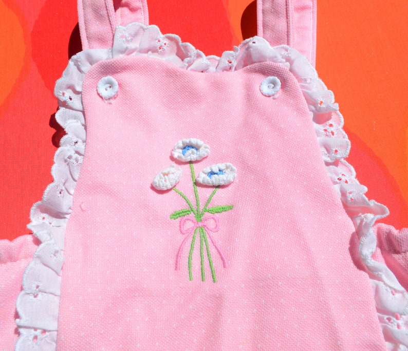 Puede incluir: Mono rosa para beb&eacute; con ribete de encaje blanco y flores bordadas. Las flores son blancas con centros azules y tallos verdes. Hay un lazo rosa en los tallos.