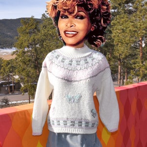 May include: White knitted sweater with a pink, gray, and blue geometric pattern. The sweater has a mock neck and long sleeves.