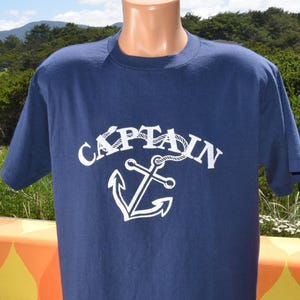 May include: Navy blue t-shirt with the word "CAPTAIN" in white, arched above a white anchor design. The shirt has short sleeves and a crew neck. The design is nautical-themed.