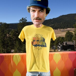 Pu&ograve; includere: Una maglietta gialla con una grafica vintage di "North Carolina" in un design a forma di sole. La maglietta &egrave; indossata da una persona che indossa un cappello da baseball blu.