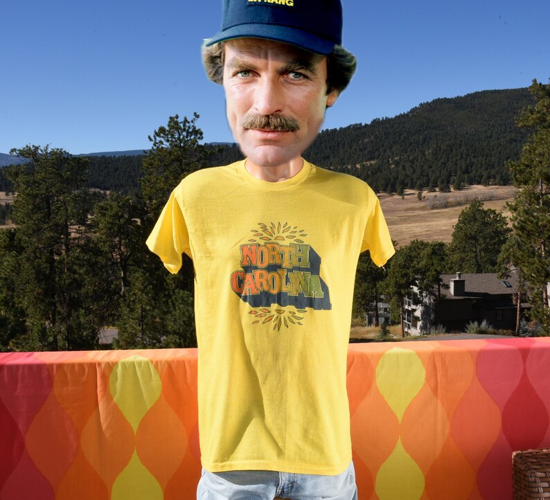 Pu&ograve; includere: Una maglietta gialla con una grafica vintage di "North Carolina" in un design a forma di sole. La maglietta &egrave; indossata da una persona che indossa un cappello da baseball blu.