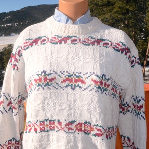 May include: White knitted sweater with a colorful geometric pattern. The sweater has a crew neck and long sleeves.