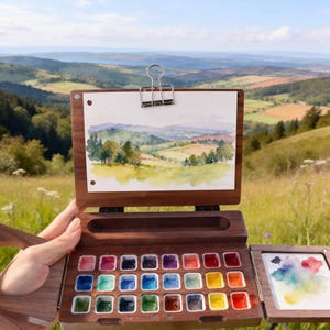 Op de afbeelding: Een houten aquarelverfdoos met een landschapsschilderij bovenaan vastgeklemd. De doos is open en onthult een palet met kleurrijke aquarelverf en een klein menggebied. De schildering toont een groen en bruin landschap met een blauwe lucht.