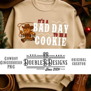 Puede incluir: Sudadera beige con la frase "It's a bad day to be a cookie" y un diseño de hombre de jengibre vaquero. Debajo, el logo de Double B Designs, "Original Creator" y "Since 2024".