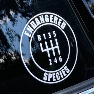 May include: A black and white sticker on a red car window. The sticker is a circle with the words "Endangered Species" around the edge. Inside the circle is a stylized gear shift pattern with the numbers 1, 3, 5, 2, 4, and 6.