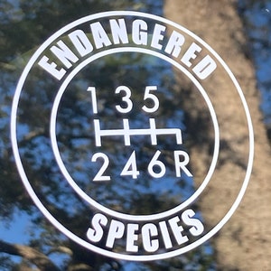 May include: A black and white sticker with the text "Endangered Species" in a circle around a gear shift pattern with the numbers 1, 3, 5, 2, 4, 6, and R.